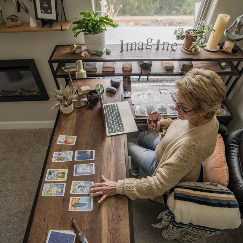 Person reading tarot cards at a cozy home workspace with laptop and plants.