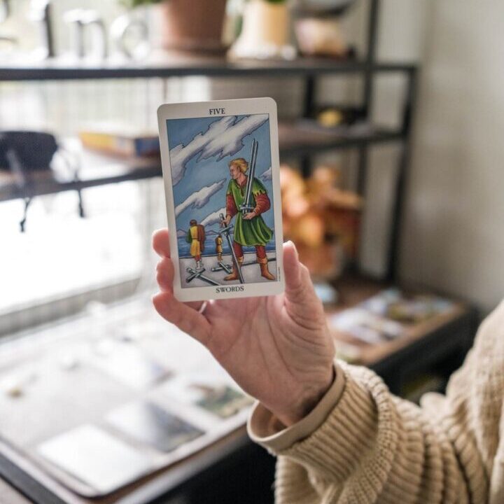 A hand holds the Six of Swords tarot card with a blurred background.